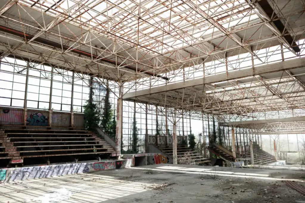 Interior de galpão industrial abandonado, com estrutura metálica exposta, escadas, grafites nas paredes e vegetação invadindo as janelas