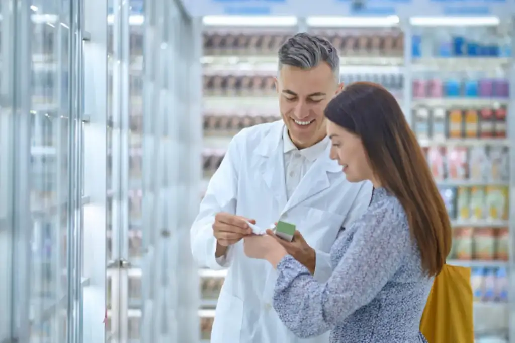 Farmacêutico de jaleco branco orienta cliente sobre medicamento em farmácia com prateleiras ao fundo