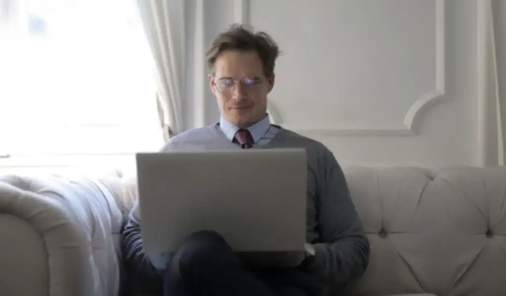 Homem de óculos e gravata sentado em sofá, sorrindo enquanto usa um laptop no colo