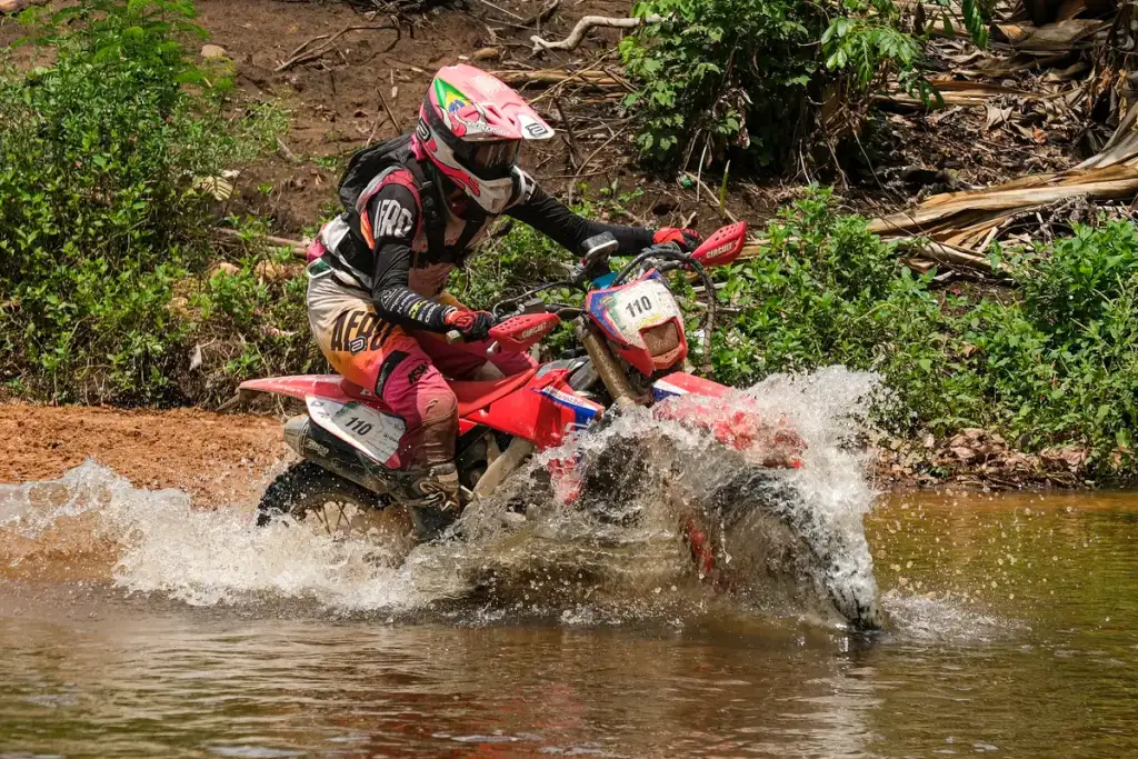 Piloto de enduro número 110 atravessa um rio, levantando muita água com a moto na travessia
