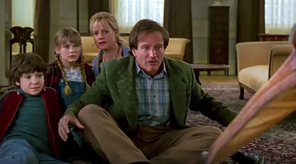 Cena do filme Jumanji com Robin Williams sentado no chão da sala, cercado por duas crianças e uma mulher, todos surpresos observando algo fora de quadro