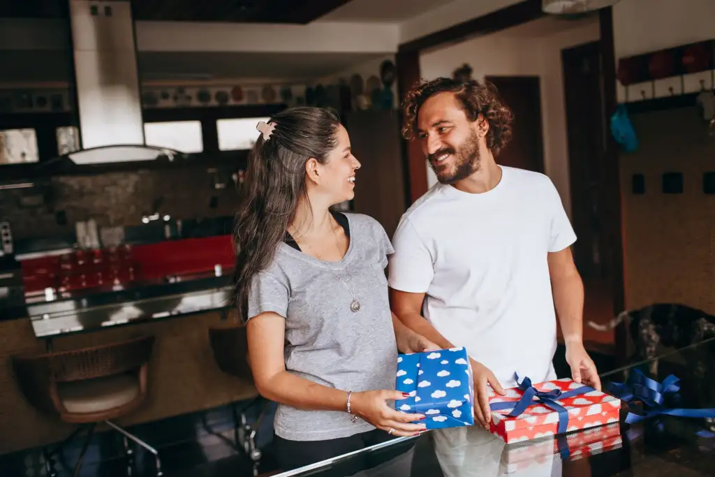 Casal sorrindo um para o outro na cozinha, segurando presentes embrulhados com laços azuis