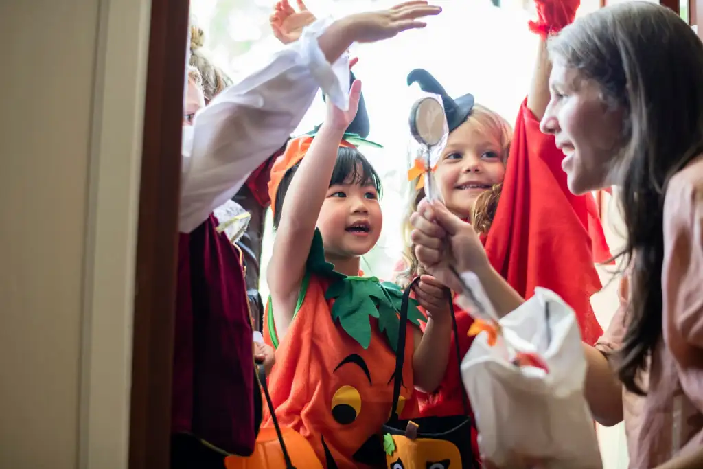 Crianças fantasiadas de Halloween, incluindo uma vestida de abóbora, recebem doces de uma mulher na porta