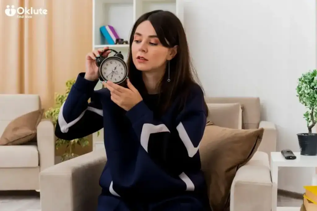 Mulher sentada em uma poltrona observa atentamente um despertador que segura na mão, em uma sala de estar