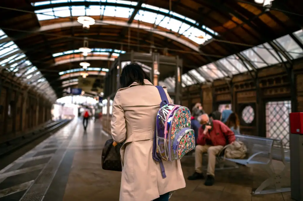 Mulher de trench coat e mochila estampada caminha na plataforma de uma estação de trem com teto arqueado