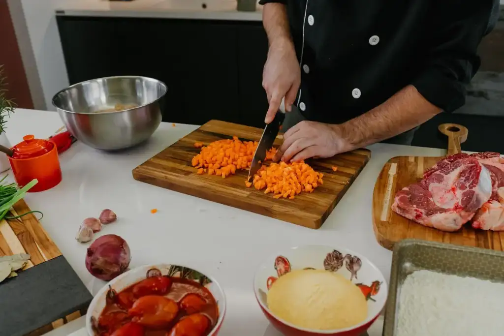 Chef picando cenoura em tábua de madeira, cercado de carne, cebola, alho e outros ingredientes na bancada