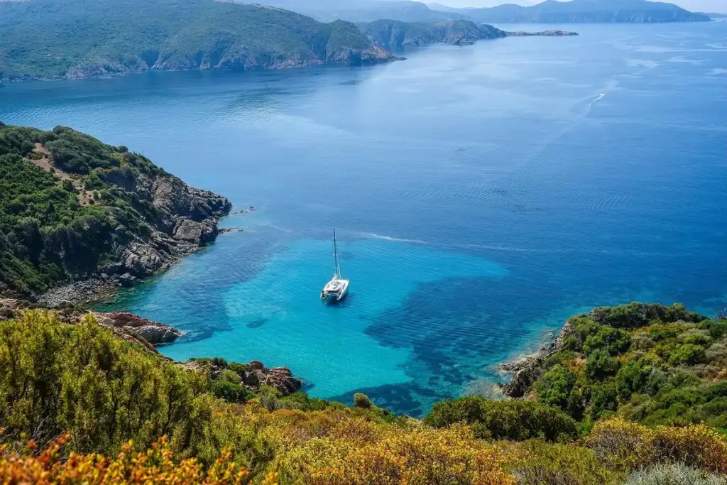 Veleiro ancorado em baía de águas turquesa cercada por costa rochosa e vegetação mediterrânea