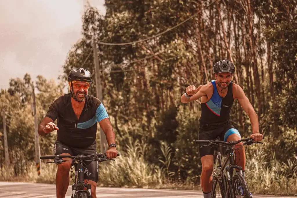 Dois ciclistas sorridentes com capacetes e roupas esportivas pedalam em estrada arborizada, comemorando com os braços erguidos
