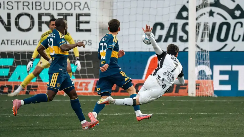 Jogador de branco chuta de bicicleta perto do gol, marcado por dois jogadores do Boca Juniors