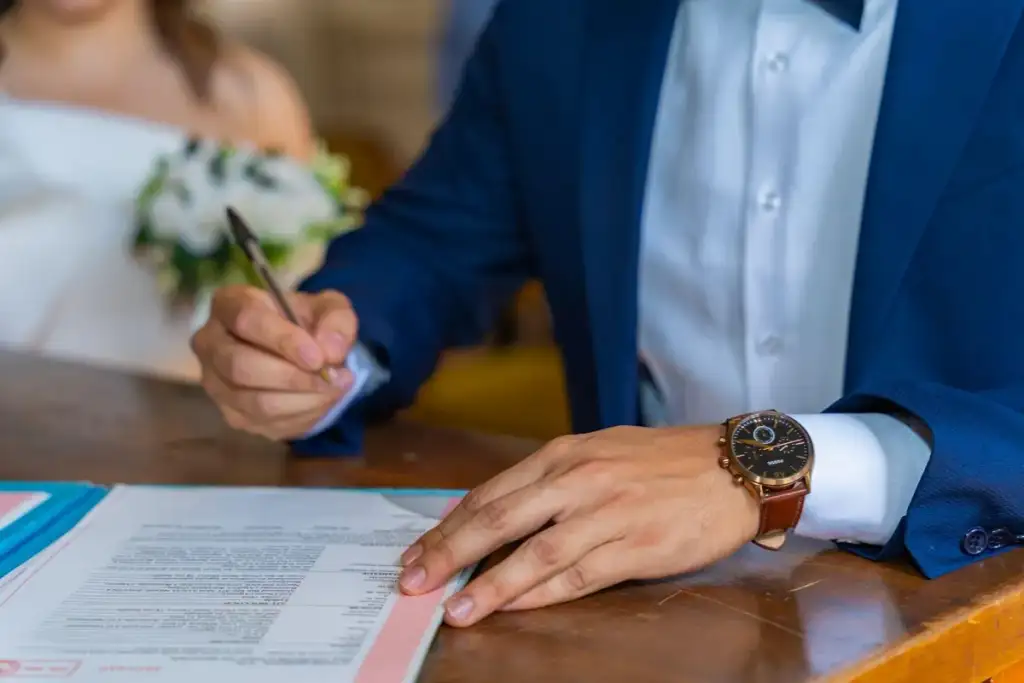Noivo de terno azul assina a certidão de casamento, com a noiva segurando buquê ao fundo