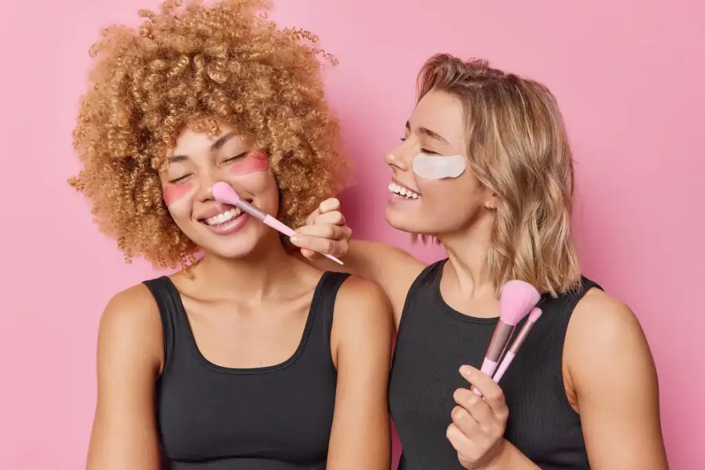 Duas mulheres sorridentes com adesivos para olheiras aplicam blush com pincel, sobre fundo rosa