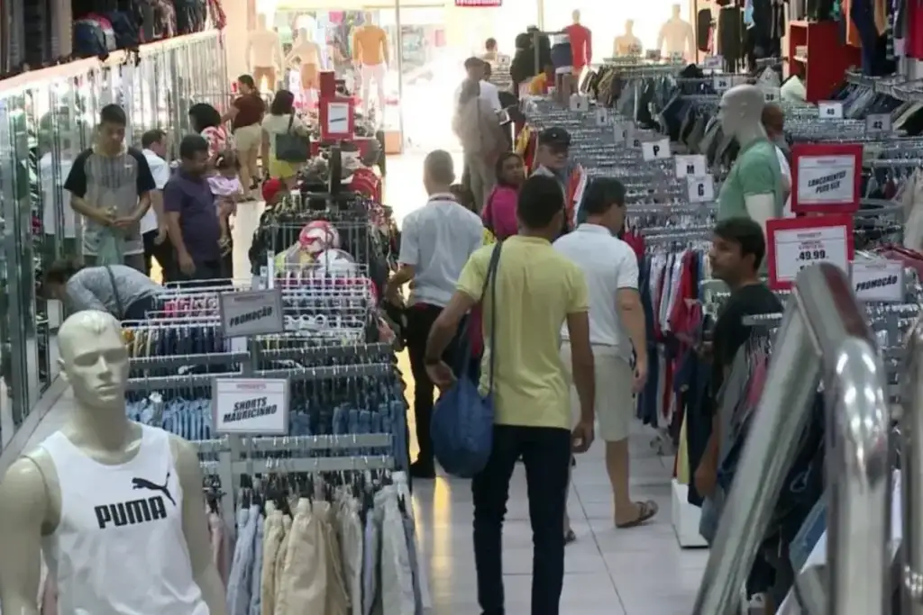 Loja de roupas movimentada com clientes circulando entre araras de roupas e cartazes de promoção