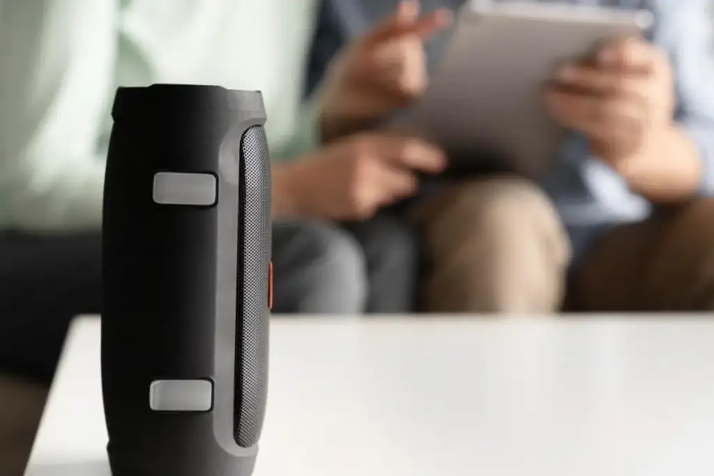 Caixa de som Bluetooth portátil preta sobre mesa, com pessoa usando tablet ao fundo desfocado