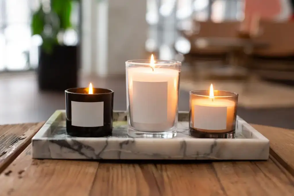 Três velas aromáticas com rótulos em branco, acesas sobre bandeja de mármore em uma mesa de madeira