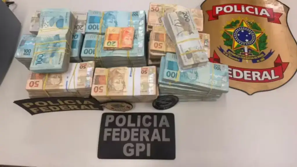 Maços de dinheiro em reais apreendidos exibidos ao lado do brasão da Polícia Federal e placa "Polícia Federal GPI