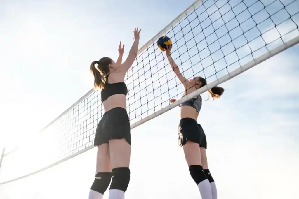 Duas jogadoras disputam a bola perto da rede durante uma partida de vôlei, uma bloqueando e outra atacando